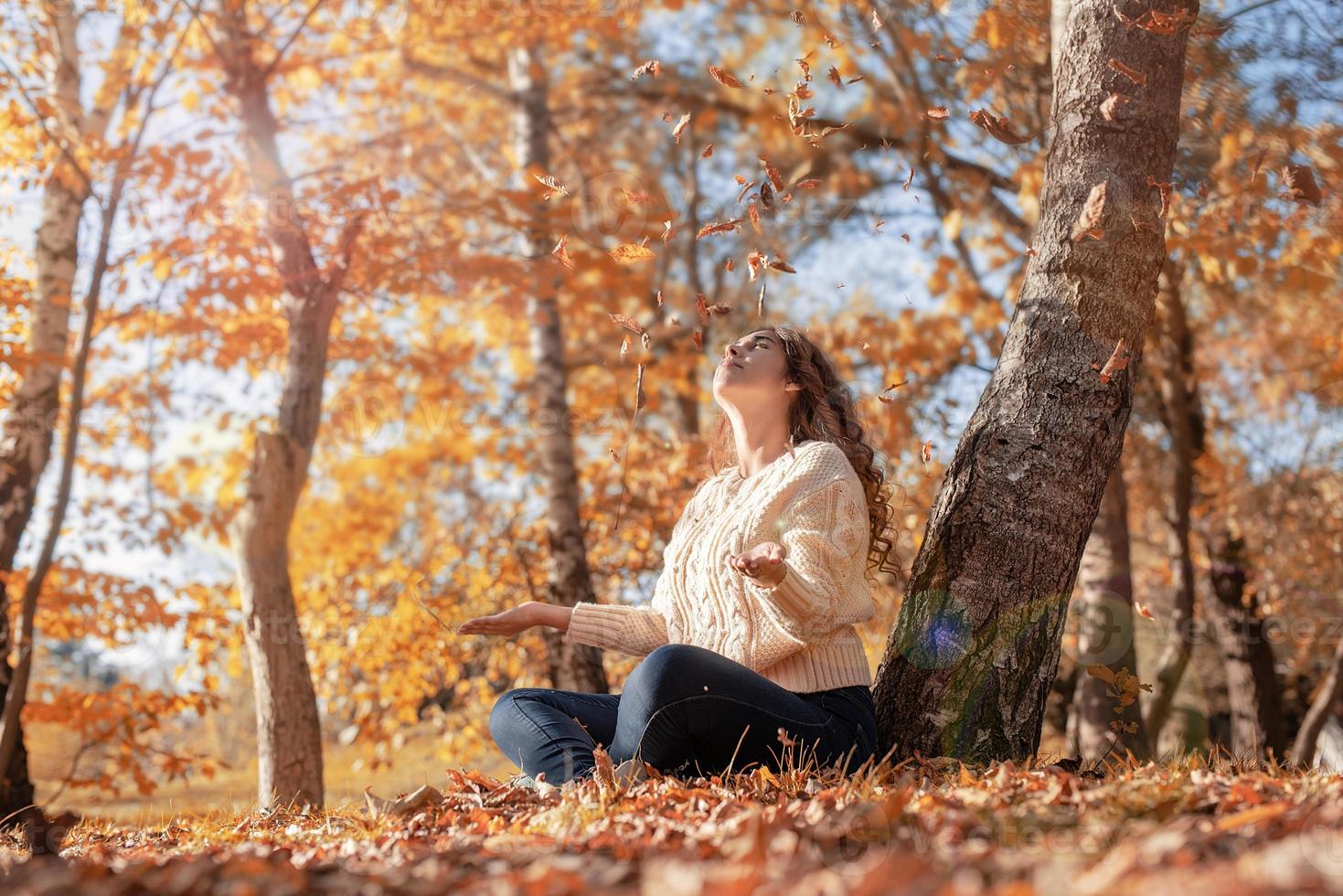 jonge vrouw die gele bladeren in de lucht gooit terwijl ze in het herfstbos zit foto