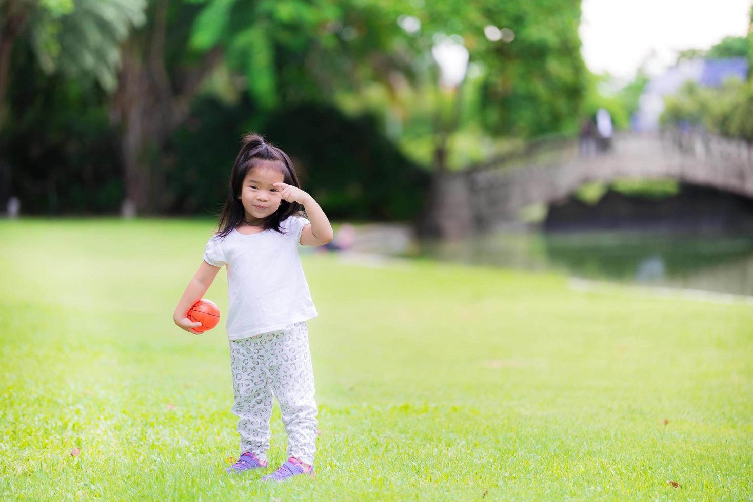 gelukkig meisje kleine oranje bal spelen in groen gras met familie. picknick met ouders op vakantie. winter in centraal thailand. fris weer. schattig 3-jarig kind, lieve glimlach, kind dat wenkbrauwen krabt foto