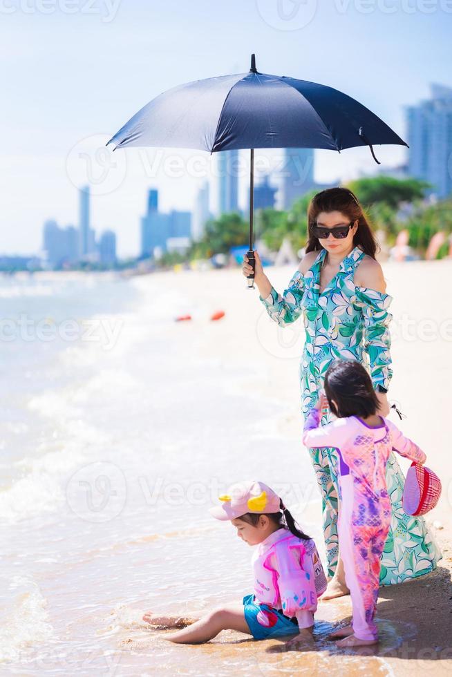 verticaal beeld. familie reizen aan zee en strand. tante en kleindochters spelen aan zee. vrouw met zwarte paraplu beschermt hete zon. zomerdag. gelukkig kind meisje draagt hoed en zittend op zand. foto