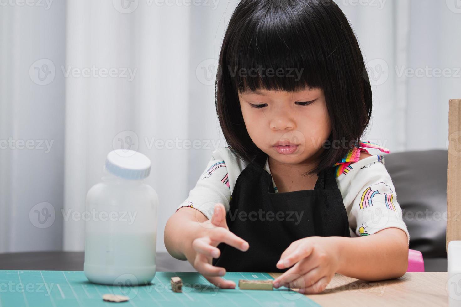 leerling meisje doet papier ambacht op homeschool. kleuterschool student lijmen. gelukkig kind dat zwarte schort draagt. baby van 4-5 jaar oud. foto