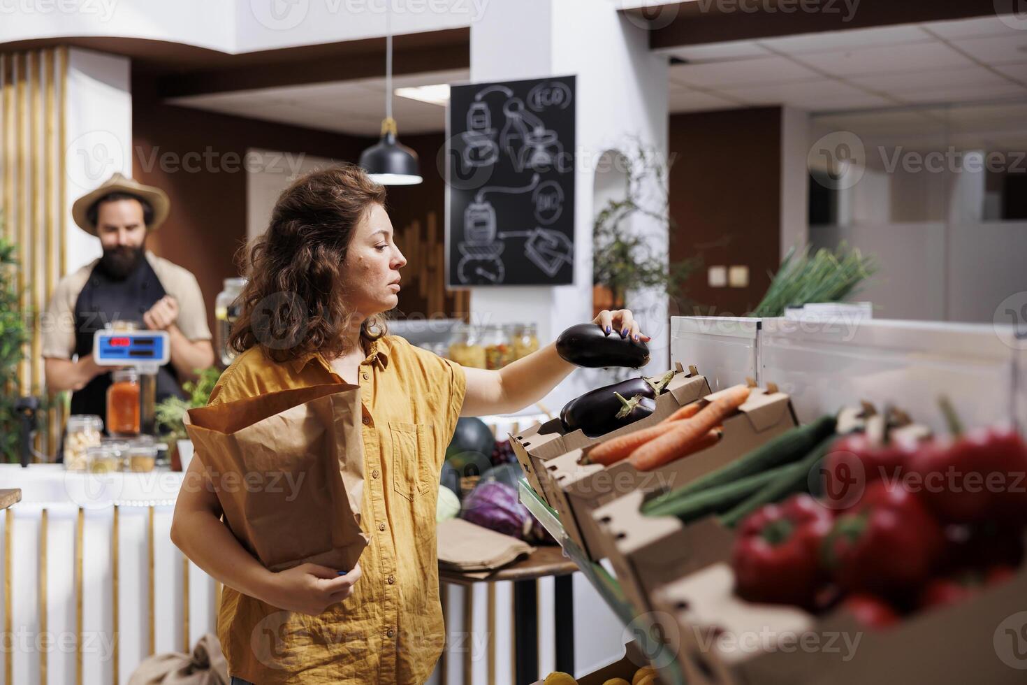 vrouw in nul verspilling winkel inkoop biologisch plaatselijk gegroeid groenten, plukken rijp aubergines. klant in lokaal kruidenier winkel op zoek naar kopen gezond voedsel, gebruik makend van papier zak naar vermijden single gebruik kunststoffen foto