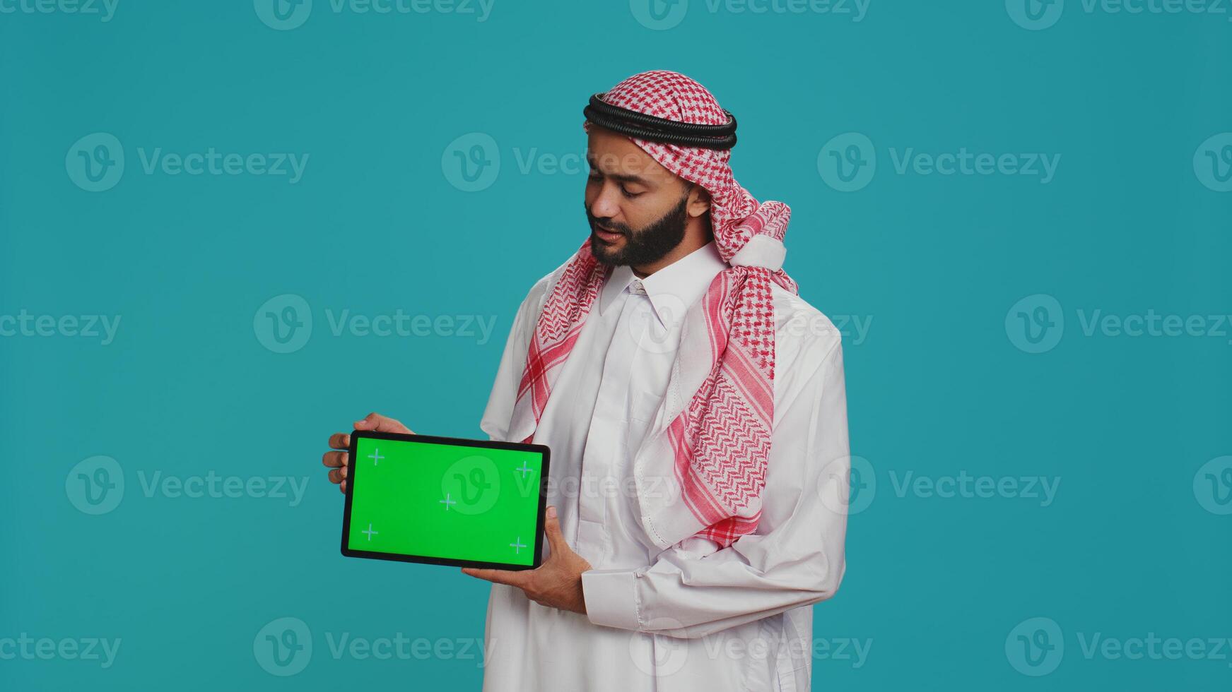 moslim Mens presenteert groene scherm tablet over- blauw achtergrond, tonen blanco copyspace Scherm. jong persoon in Arabisch kleding Holding apparaatje met geïsoleerd Chroma sleutel mockup sjabloon. foto