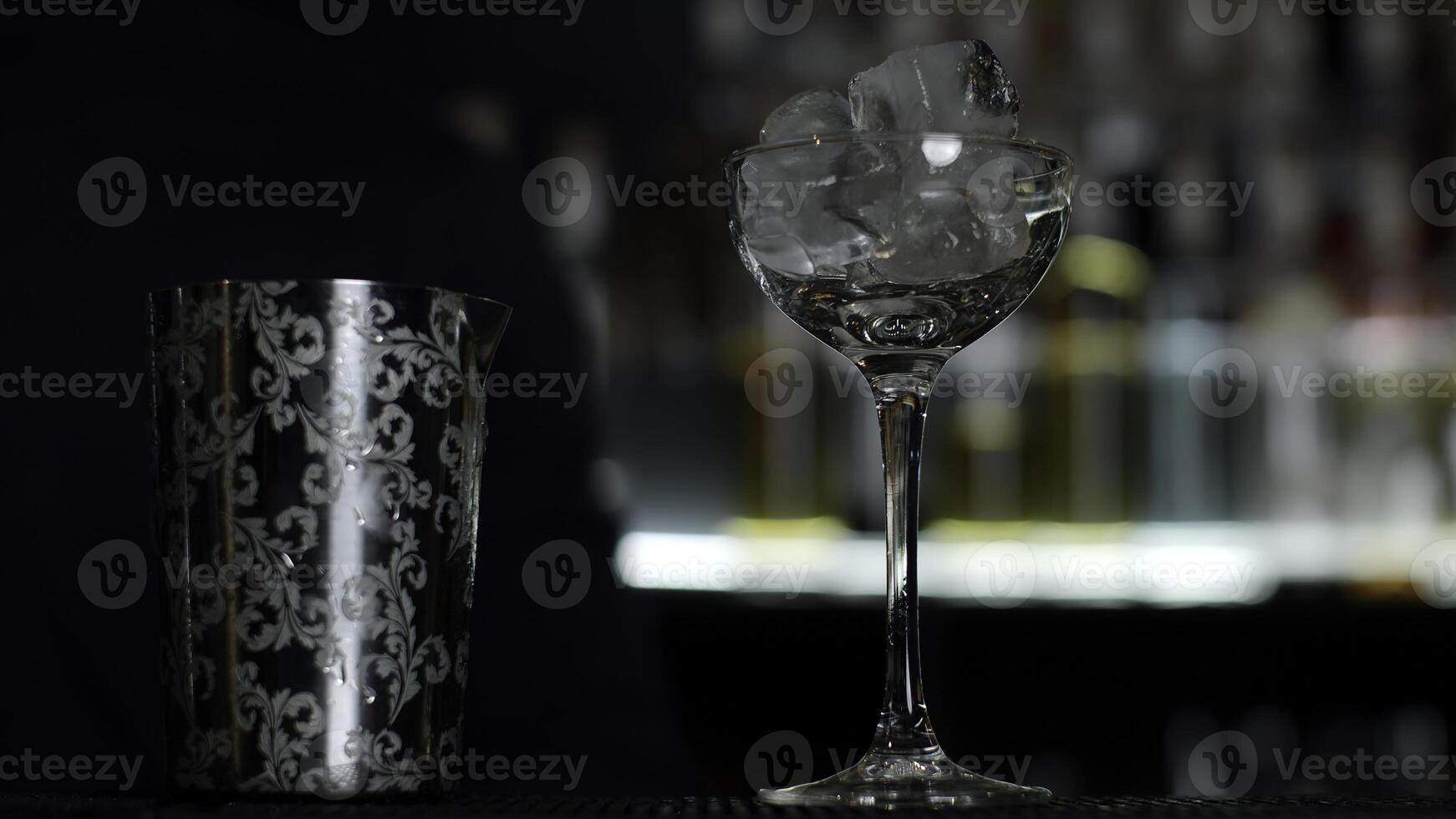 dichtbij omhoog van een barman zetten ijs kubussen in de klein leeg cocktail glas. media. details van maken een drinken met wazig flessen van alcohol Aan de achtergrond. foto