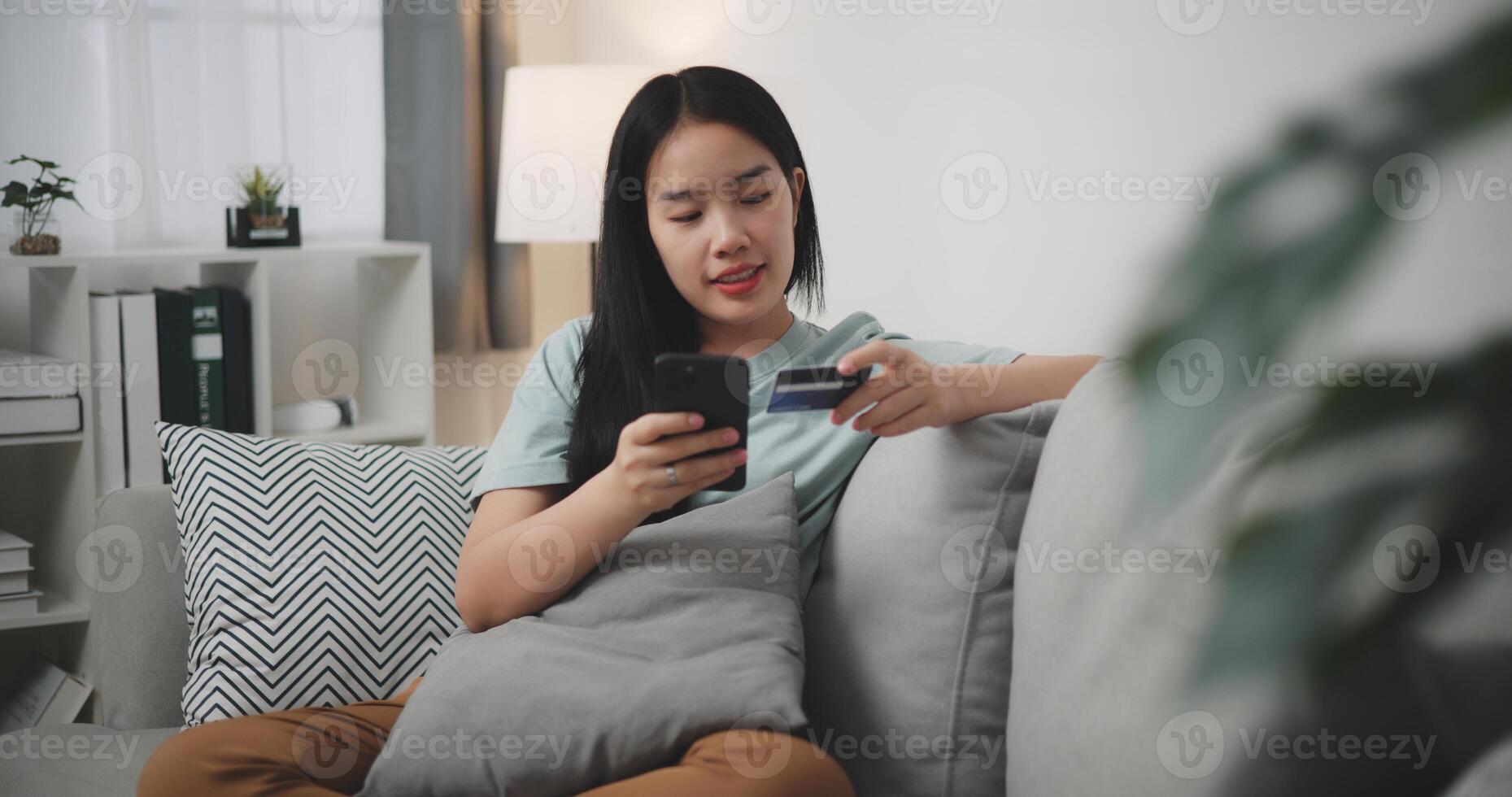selectief focus, jong Aziatisch vrouw zittend Aan sofa Holding credit kaart maken online betaling voor aankoop in web op te slaan gebruik makend van smartphone. foto