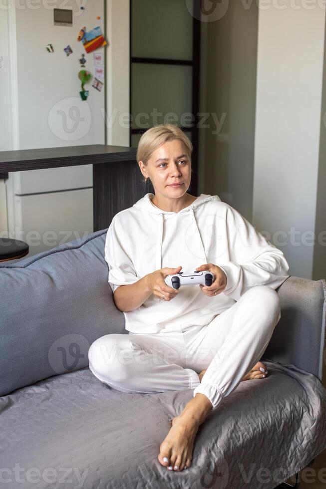 volwassen vrouw in wit zittend Aan een bankstel spelen video spellen. foto