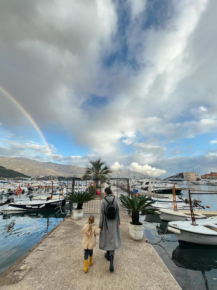 budva, Montenegro - 25 december 2022. mam met een weinig meisje wandelen langs de pier tegen de backdrop van een regenboog over- de bergen foto