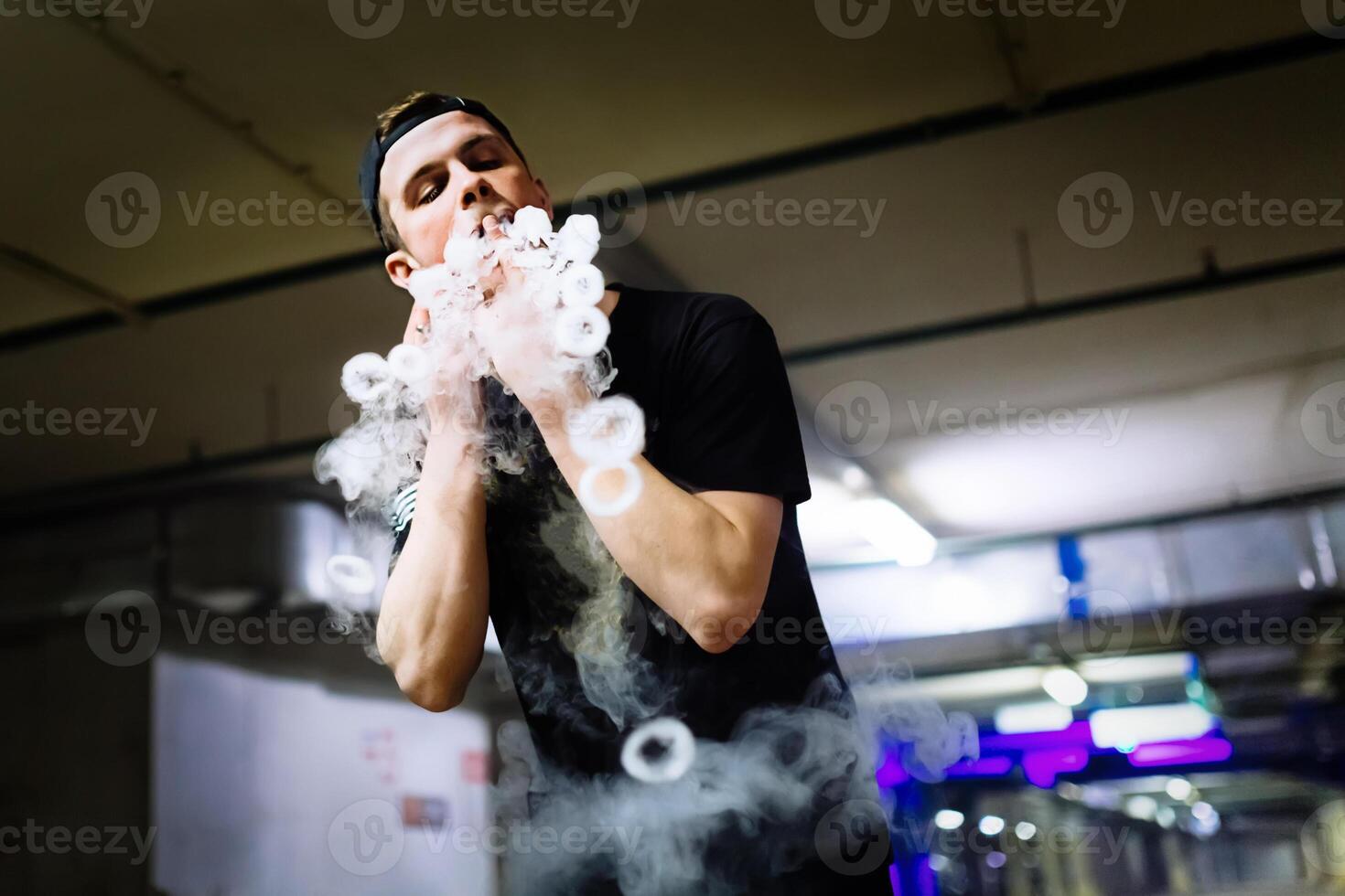 Mens in pet rook een elektronisch sigaret en releases wolken van damp het uitvoeren van divers soort van vapen trucs foto