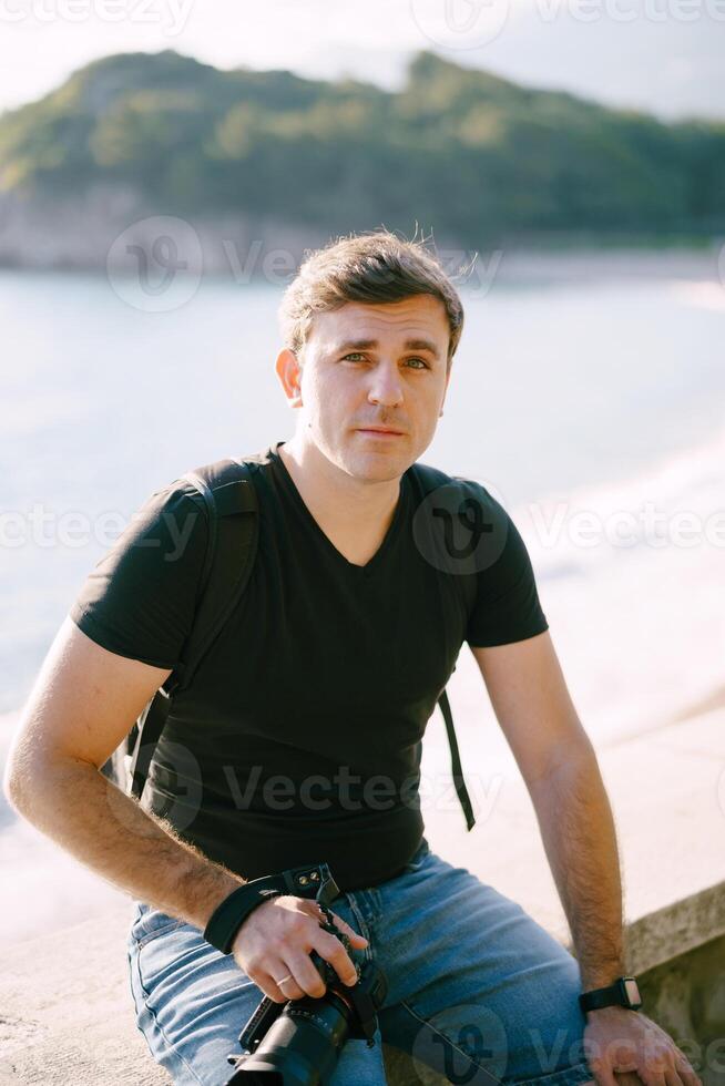 jong Mens met een camera in zijn hand- zit Aan een hek door de zee foto