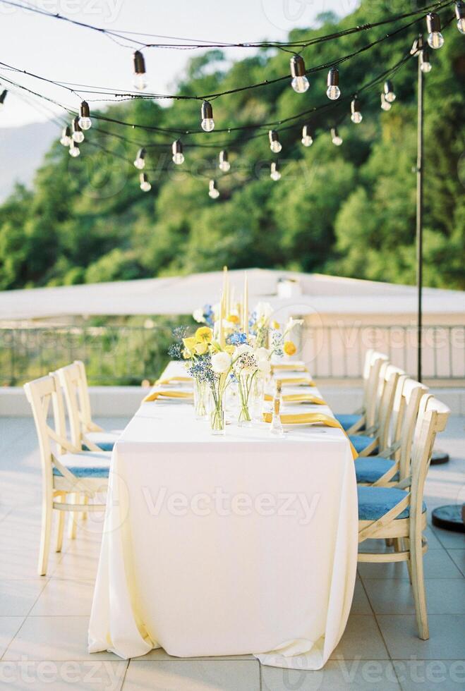 lang feestelijk tafel met boeketten van bloemen Aan de terras met hangende slingers van licht bollen foto