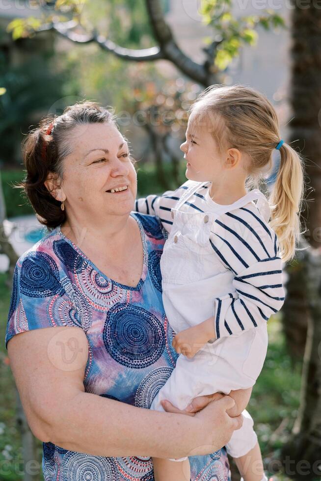 weinig meisje op zoek Bij glimlachen grootmoeder terwijl zittend in haar armen in de tuin foto