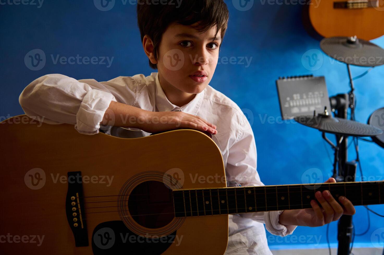 knap tiener- jongen spelen gitaar in de modern muziek- studio, op zoek vol vertrouwen Bij camera, zittend tegen een blauw muur met elektrisch trommels. mensen. muziek. hobby's en vrije tijd foto