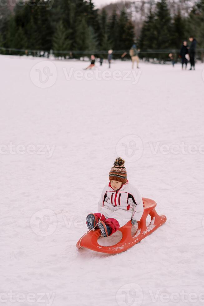 klein kind in overall zit Aan een slee Holding de handvatten Aan een besneeuwd heuvel foto