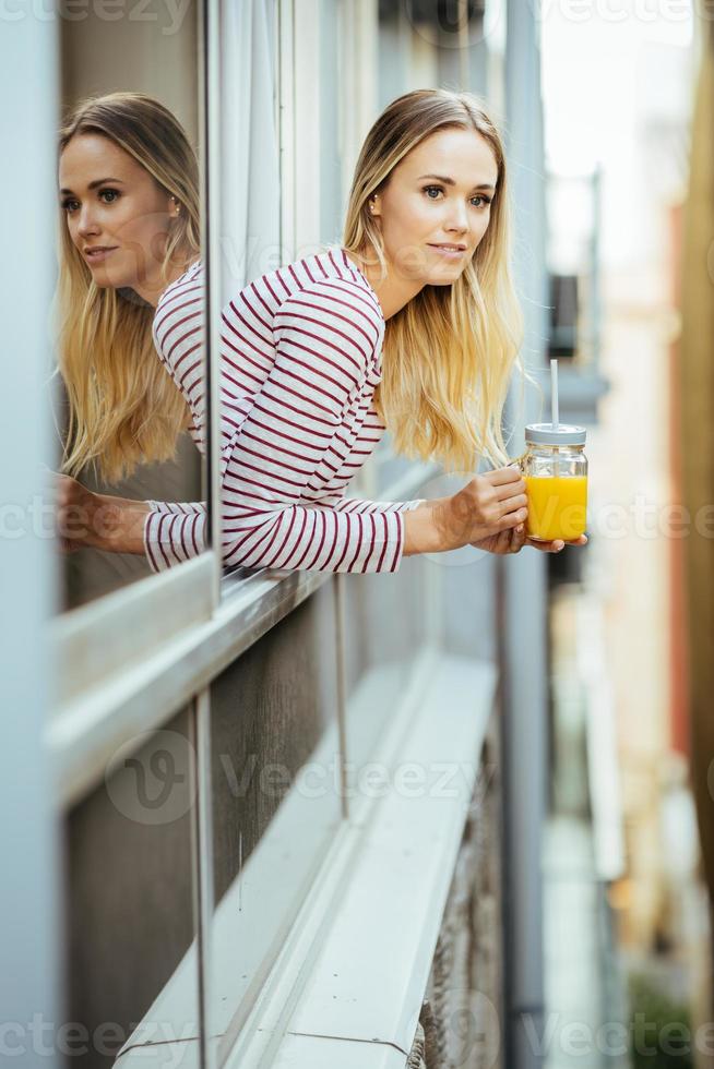 jonge vrouw die een glas natuurlijk sinaasappelsap drinkt, leunend uit het raam van haar huis. foto