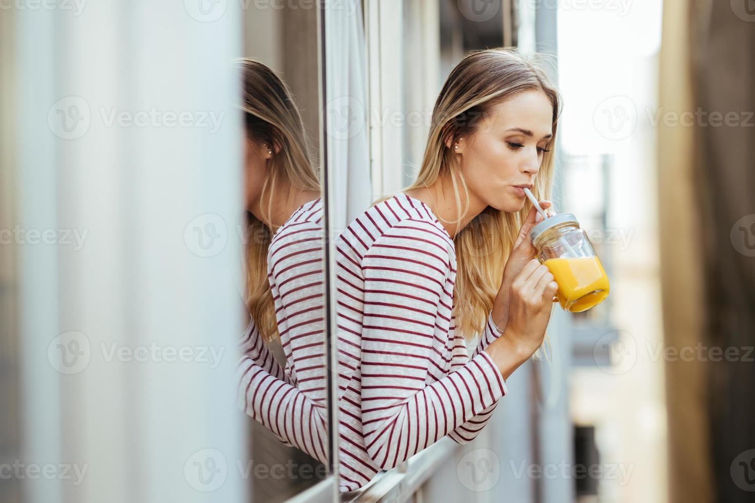 jonge vrouw die een glas natuurlijk sinaasappelsap drinkt, leunend uit het raam van haar huis. foto