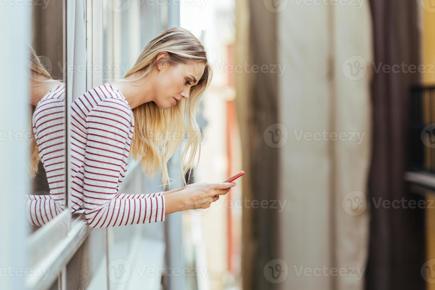 aantrekkelijke vrouw leunend uit het raam van haar huis met behulp van een smartphone. foto