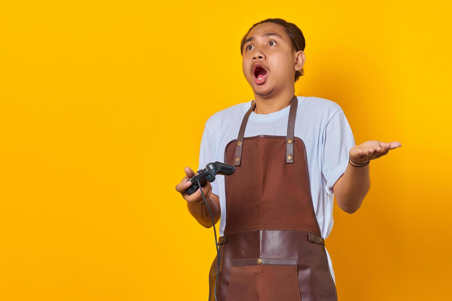 portret van een verraste aziatische jonge man met een schort met een gamecontroller met open mond wijzend op een lege ruimte met de handpalm foto