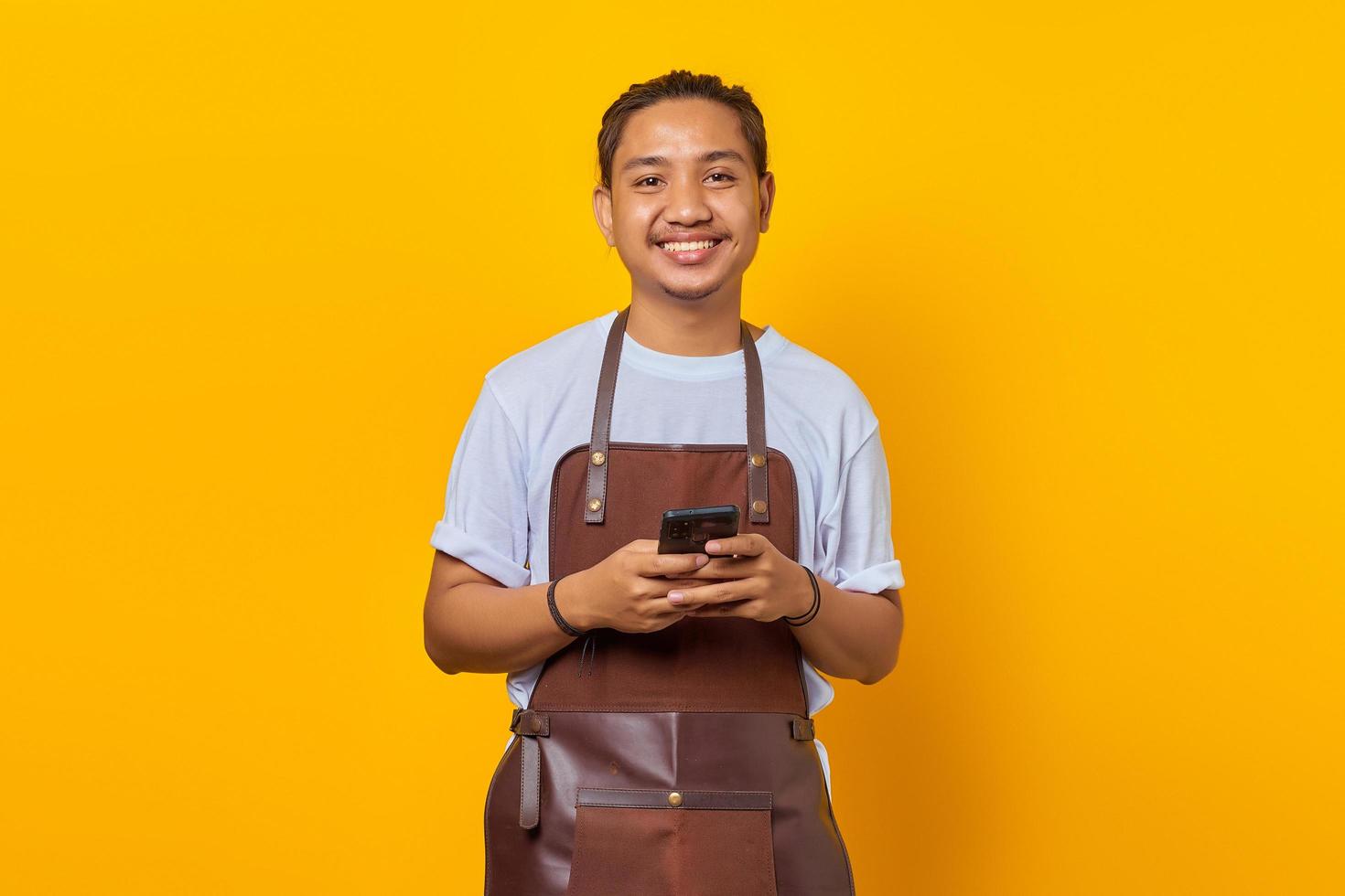 portret van een jonge aziatische man met een schort die er gelukkig uitziet met een smartphone op een gele achtergrond foto