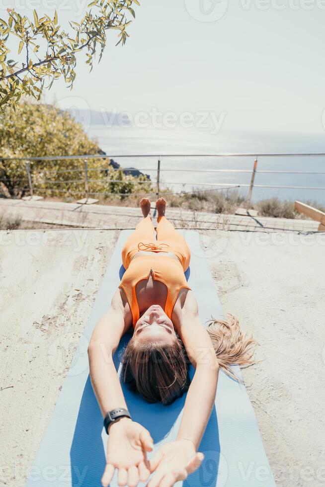 geschiktheid vrouw zee. gelukkig midden- oud vrouw in oranje sportkleding opdrachten ochtend- buitenshuis Aan yoga mat met laptop in park over- oceaan strand. vrouw geschiktheid pilates yoga routine. gezond levensstijl. foto