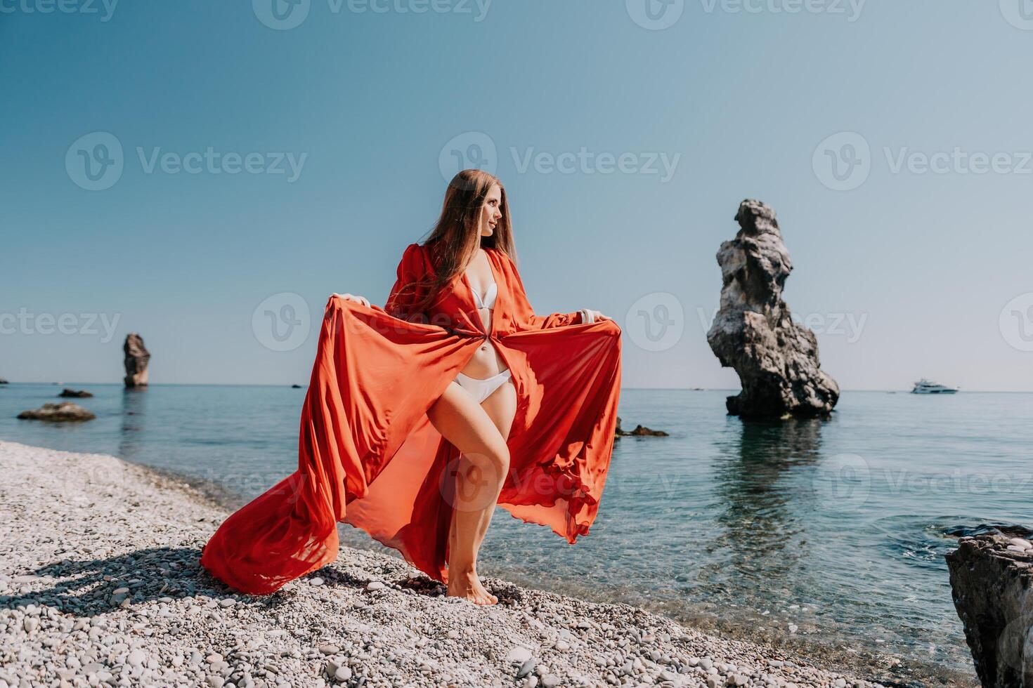 vrouw reizen zee. gelukkig toerist in rood jurk genieten nemen afbeelding buitenshuis voor herinneringen. vrouw reiziger poseren Aan de rots Bij zee baai omringd door vulkanisch bergen, sharing reizen avontuur reis foto