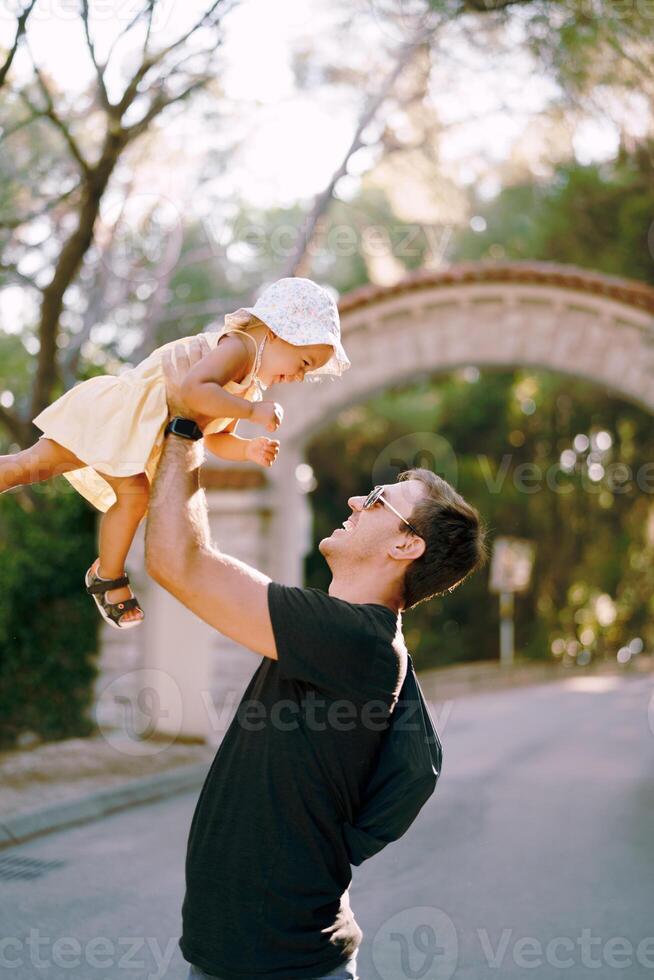 glimlachen vader liften een weinig meisje omhoog hoog in zijn armen in de buurt de park poort. kant visie foto