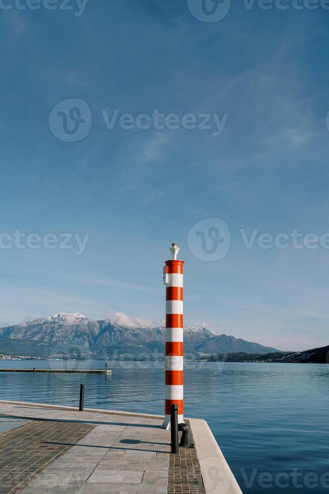 klein gestreept rood en wit vuurtoren Aan een pier door de zee tegen de backdrop van bergen foto