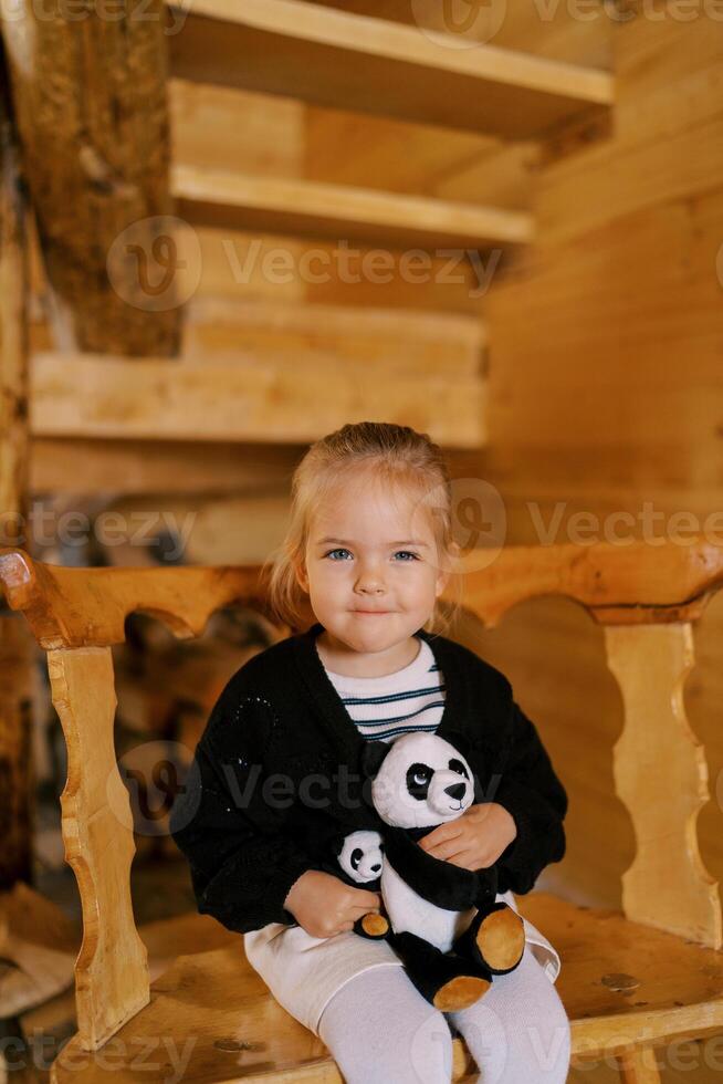 weinig glimlachen meisje zittend met zacht speelgoed- panda's Aan een houten stoel in de kamer foto