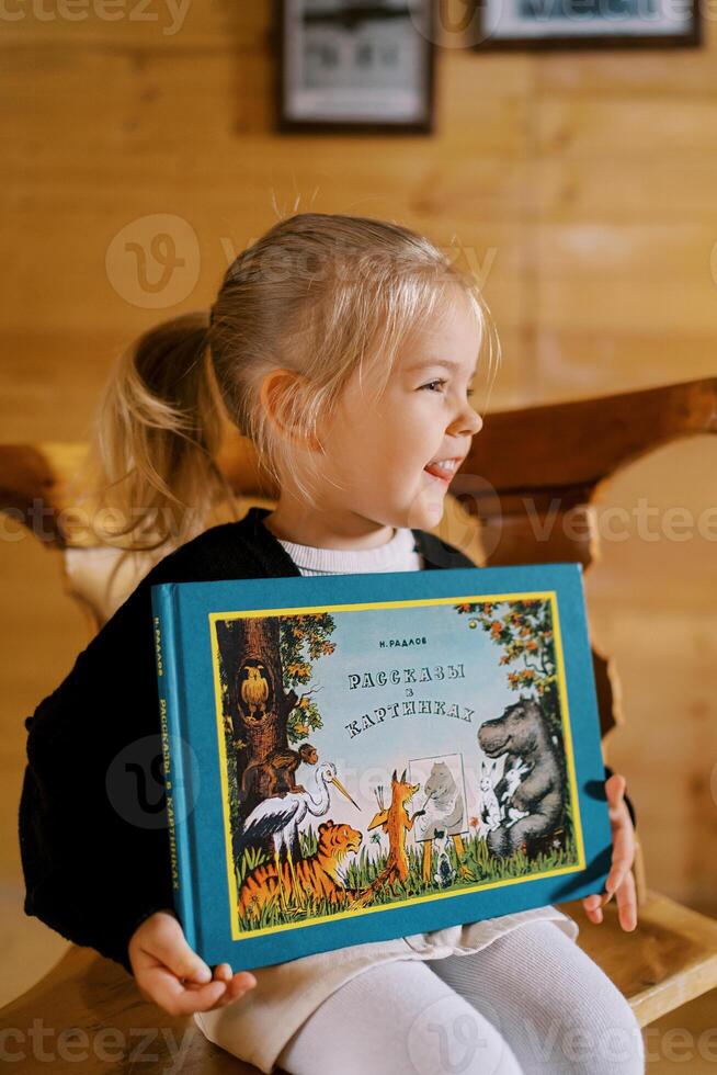 weinig glimlachen meisje zit Aan een stoel met een kleurrijk boek in haar handen. titel. verhalen in afbeeldingen foto