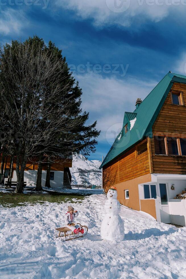weinig meisje eet van een bord zittend Aan een slee in de buurt een sneeuwman De volgende naar een houten huis foto