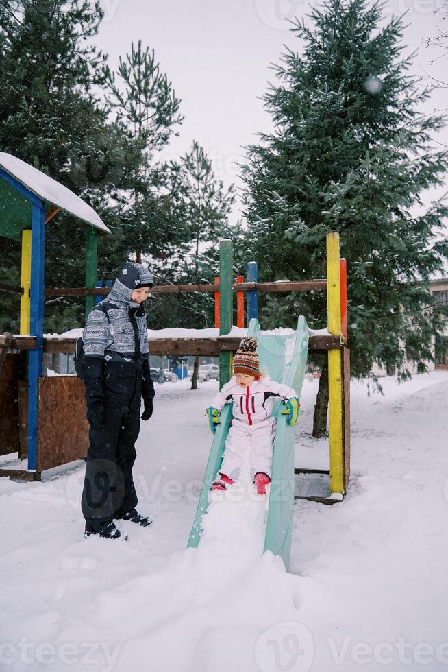 mam looks Bij een weinig meisje glijden naar beneden een met sneeuw bedekt kleurrijk glijbaan foto