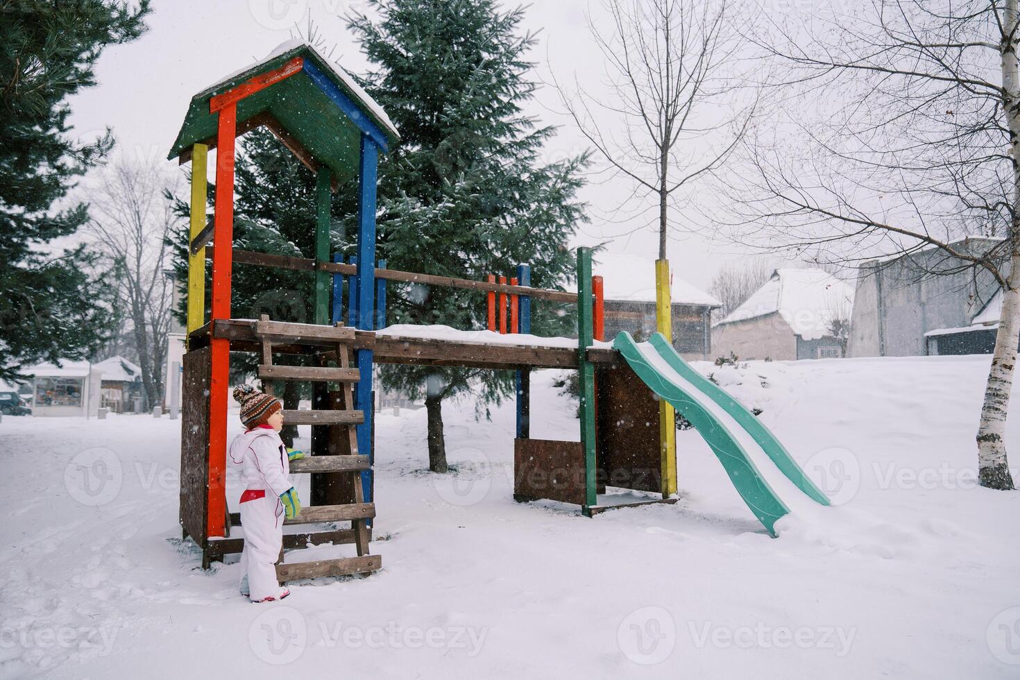 weinig meisje staat in de buurt een met sneeuw bedekt kleurrijk glijbaan tussen de bomen en looks naar de kant foto