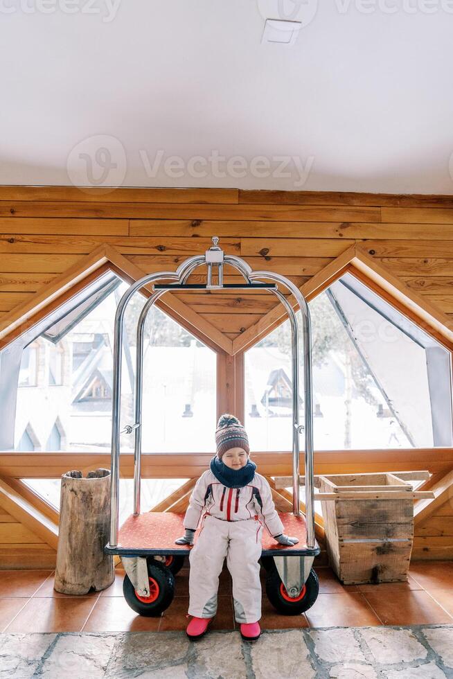 weinig meisje in een ski pak zit Aan een hotel bagage kar in de lobby foto