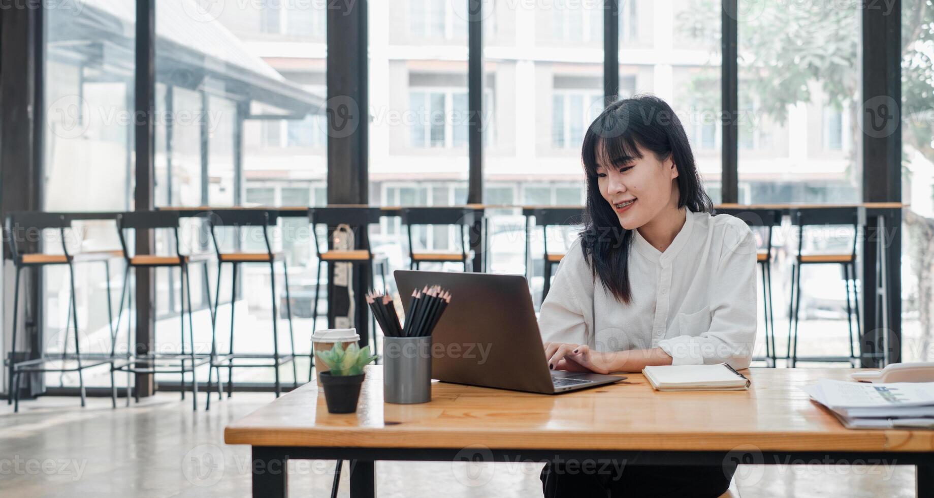 glimlachen Aziatisch vrouw gefocust Aan haar laptop werk in een helder cafe milieu met koffie en schrijfbehoeften Aan de tafel. foto