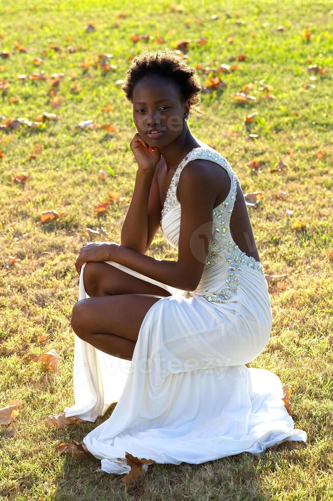 jong slank zwart vrouw in wit jurk hurken Aan gras foto
