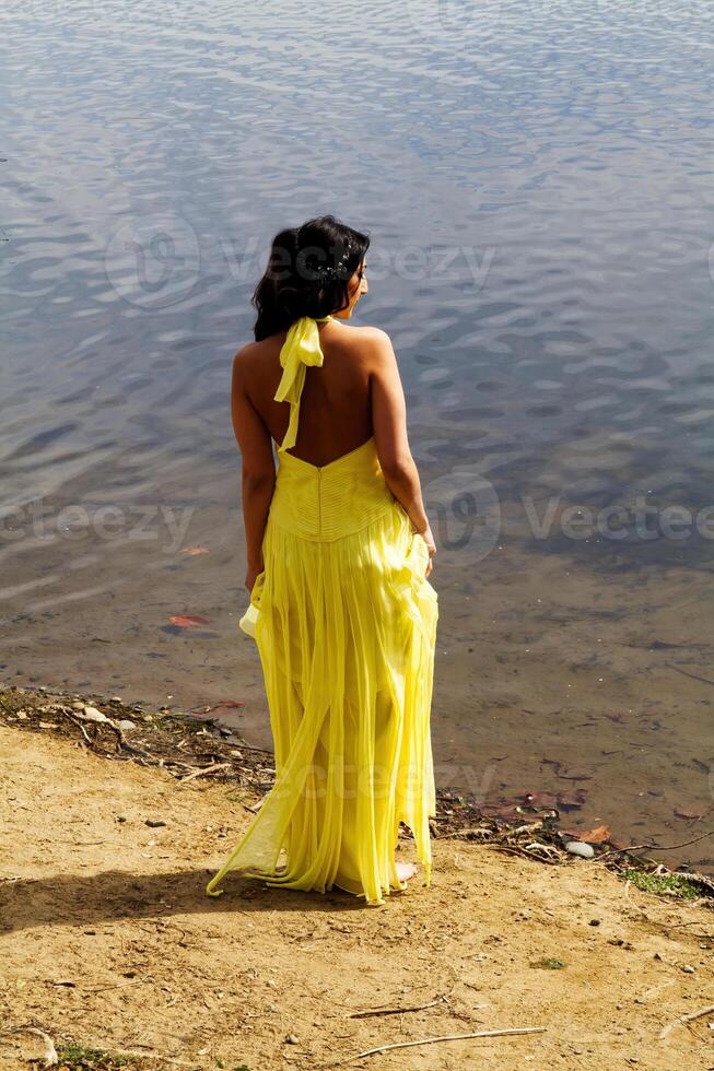 vrouw staand Aan rivier- bank op zoek Bij water in geel jurk foto