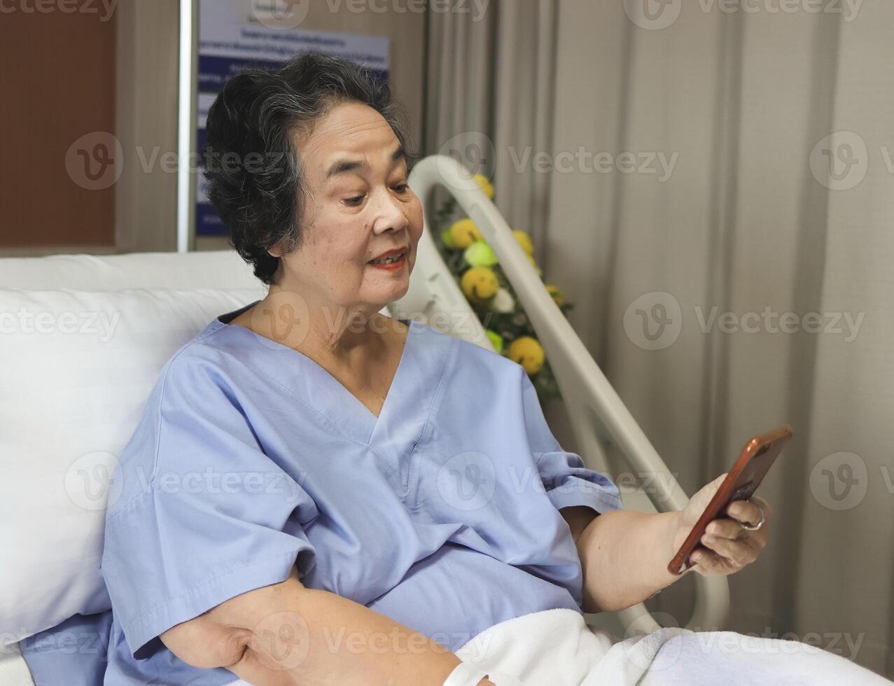 vrolijk Aziatisch senior vrouw geduldig zittend in ziekenhuis bed, glimlachen en op zoek Bij mobiel telefoon, video roeping naar haar familie. ouderen Gezondheid concept. foto