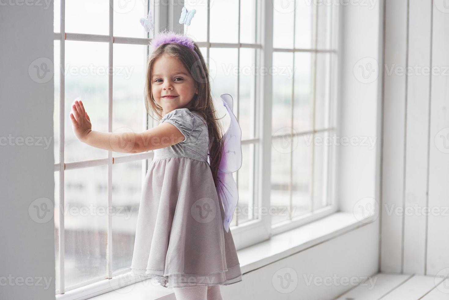 het glas aanraken. leuk klein meisje in het sprookje draagt dat bij de ramen staat en in de camera kijkt foto