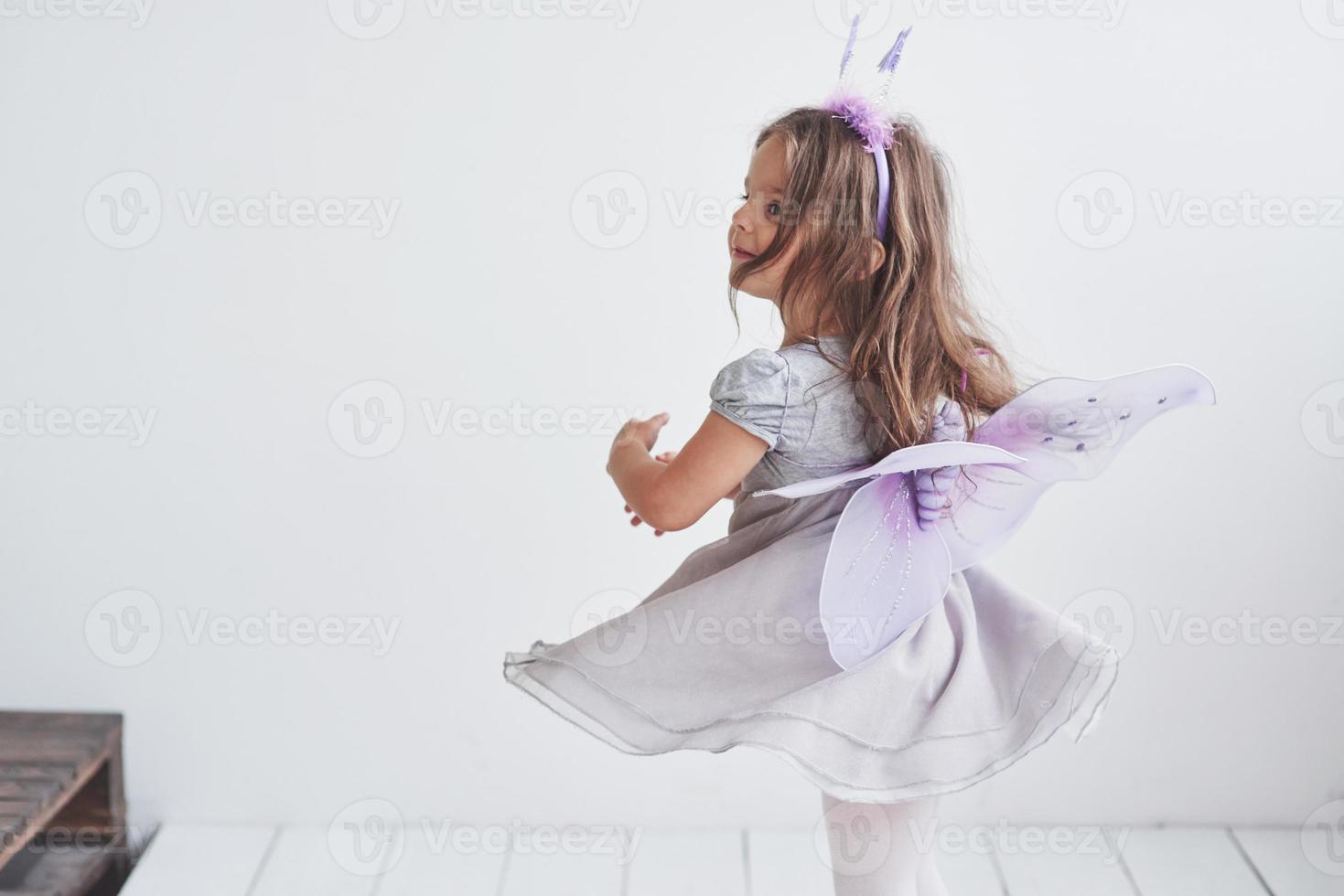 zich omdraaien en proberen te vliegen. mooi klein meisje in het sprookjeskostuum dat in de kamer staat met een witte achtergrond foto