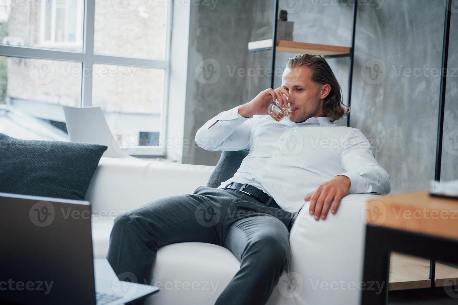 zelfverzekerde jonge man in formele kleding die voor een notebook zit en whisky drinkt foto