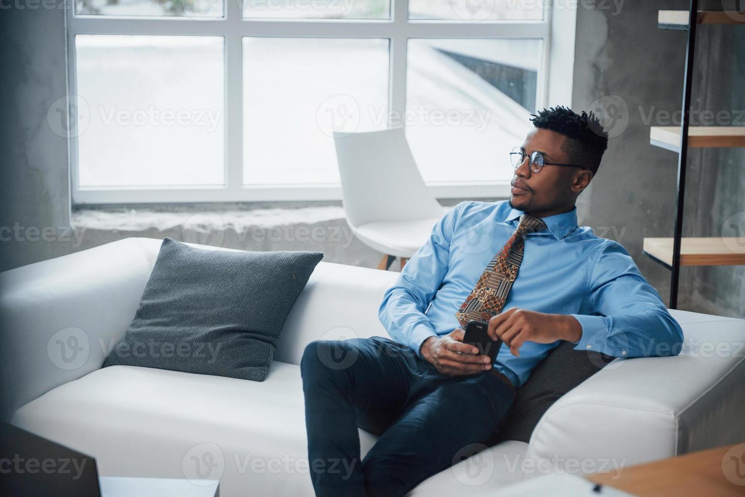 nadenkende blik van de bril. Afro-Amerikaanse man zit op de bank en kijkt naar zijn smartphone foto