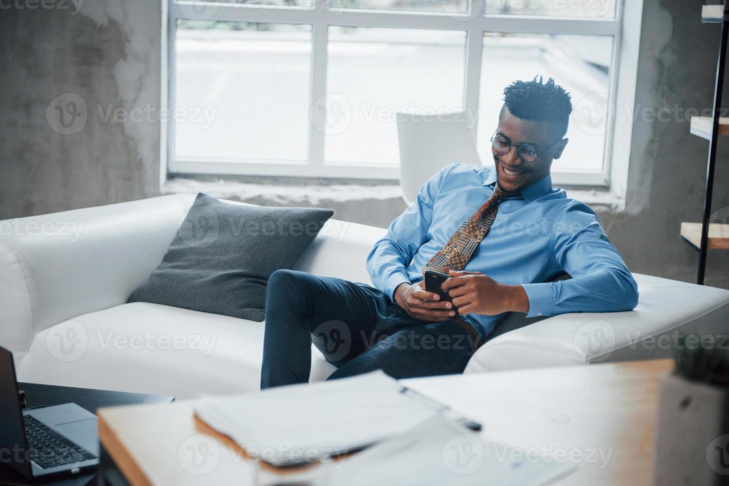 lachende Afro-Amerikaanse man zittend op de bank en kijkend naar zijn smartphone foto