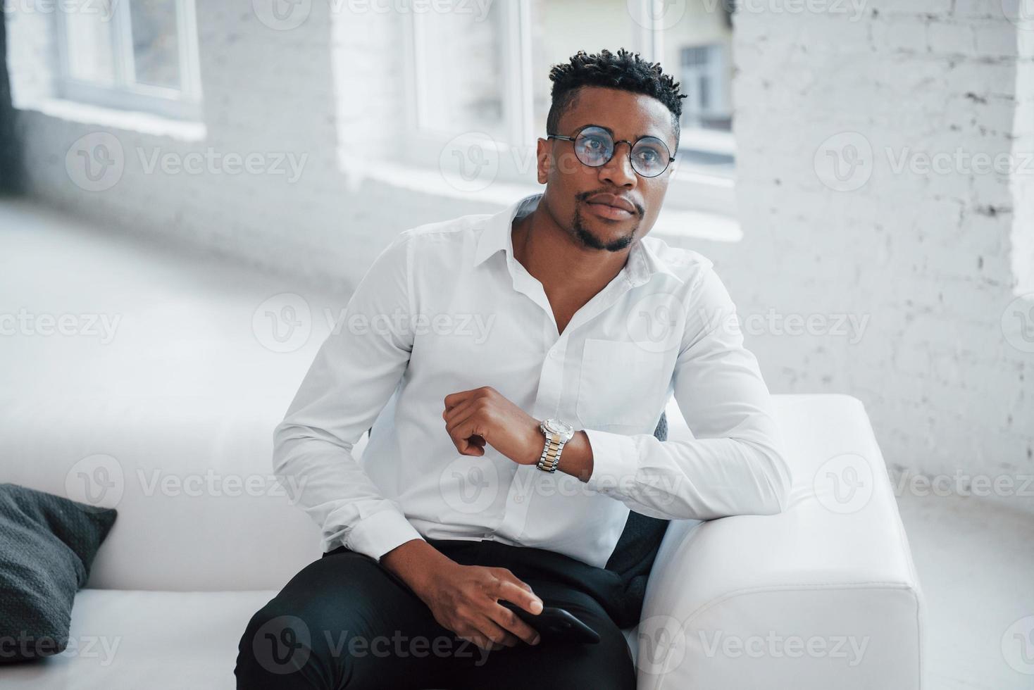 dromerige blik. stijlvolle Afro-Amerikaanse man in klassieke kleding en bril zit op het bed foto