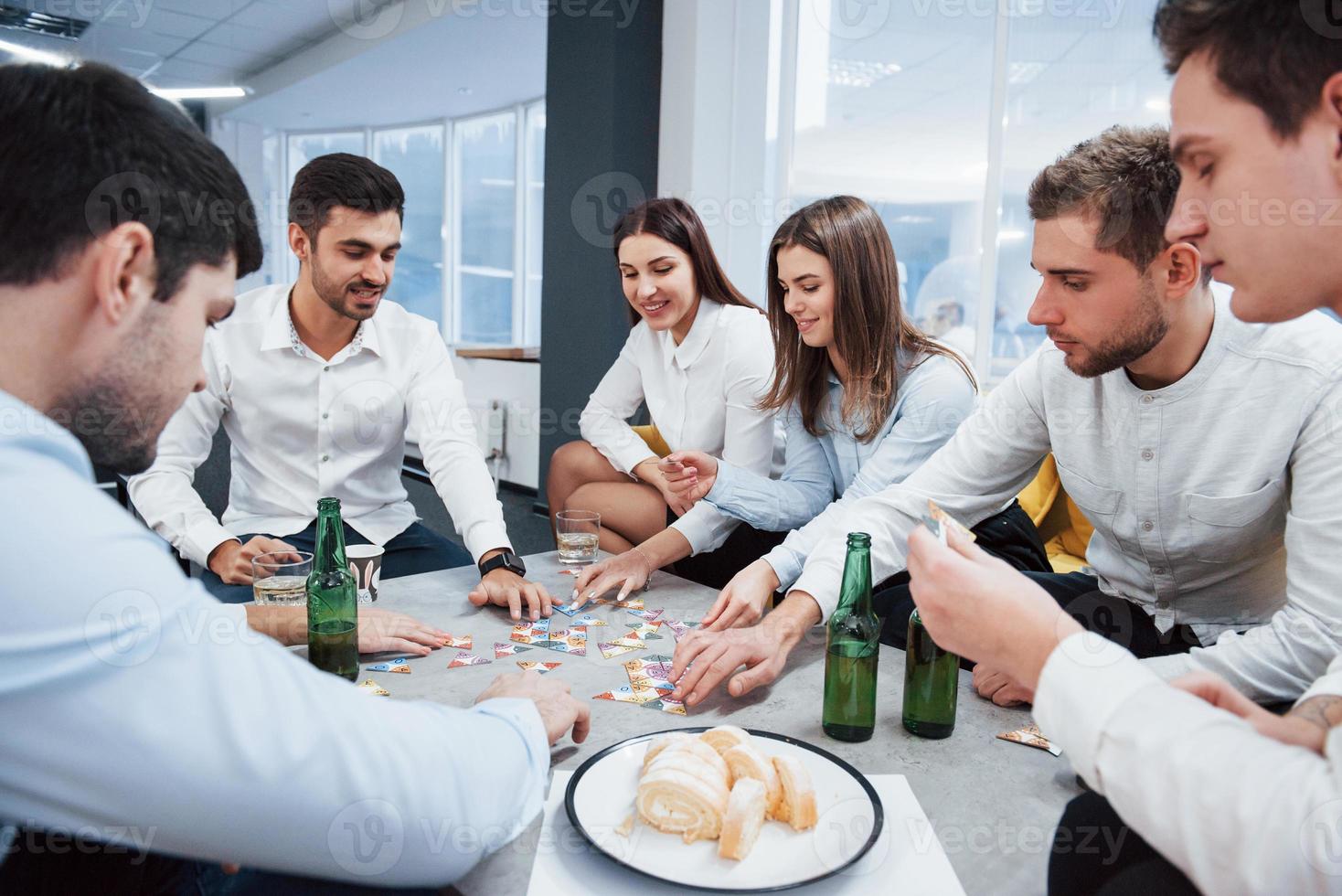 vrolijke stemming. ontspannen met spel. succesvolle deal vieren. jonge kantoormedewerkers zitten bij de tafel met alcohol foto