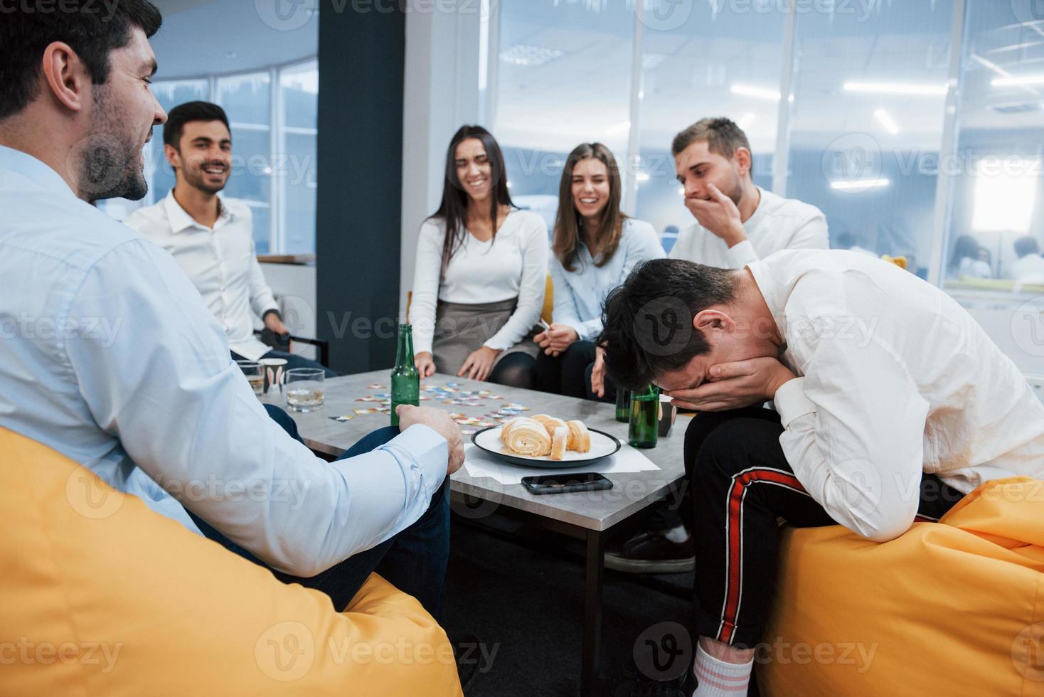 man lachen over zijn verlies met handen op het gezicht. succesvolle deal vieren. jonge kantoormedewerkers zitten bij de tafel met alcohol foto