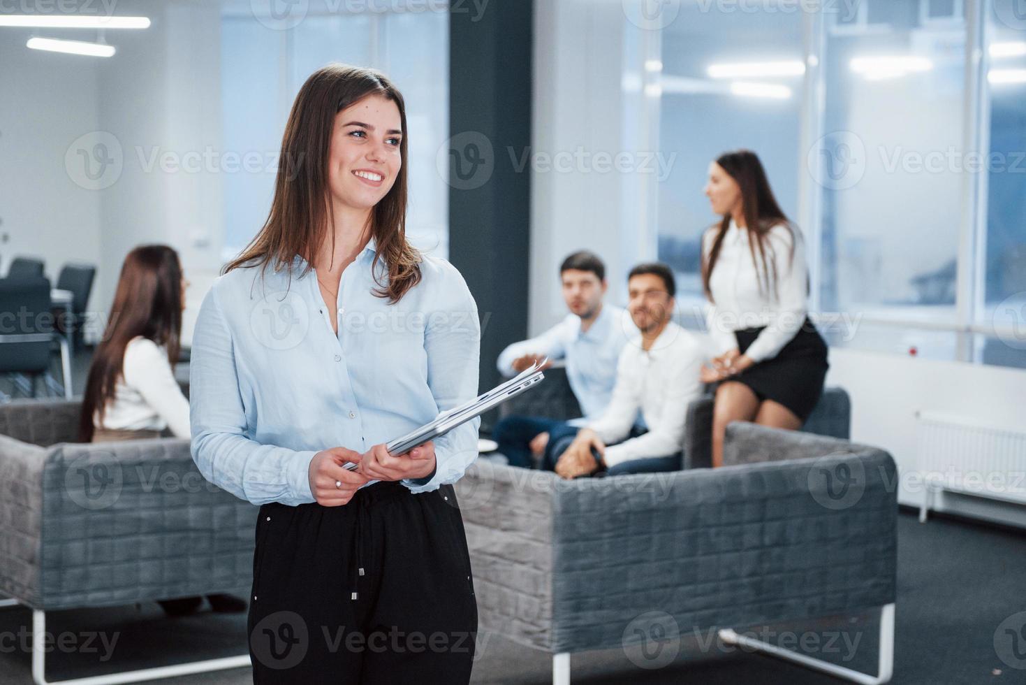 glimlachen en kijken naar de kant. portret van een jong meisje staat op kantoor met medewerkers op de achtergrond foto