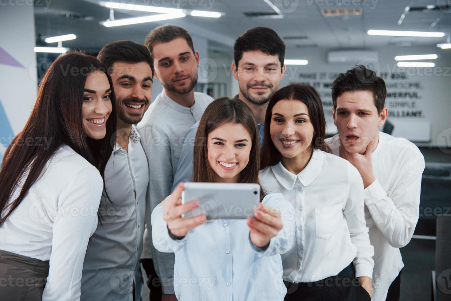 meisje houdt zilverkleurige telefoon vast. jong team selfie maken in klassieke kleding in het moderne goed verlichte kantoor foto