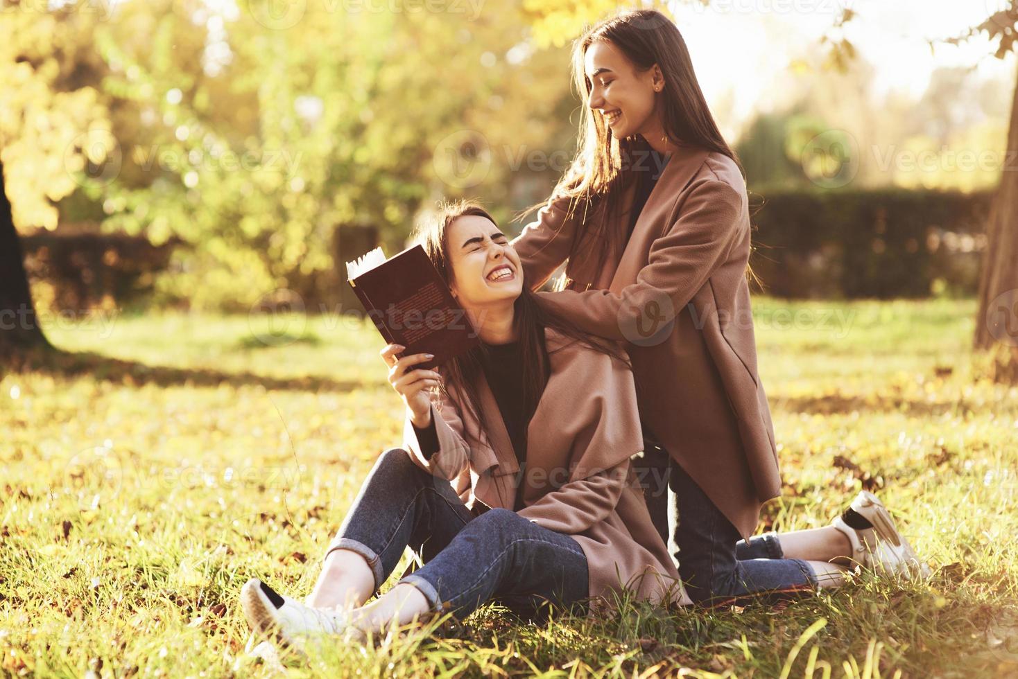 lachende brunette tweelingmeisjes die op het gras zitten. een van hen houdt een bruin boek vast, een andere leunt op de schouders van de zus met haar handen vanaf de achterkant in het herfstzonnige park op een wazige achtergrond foto