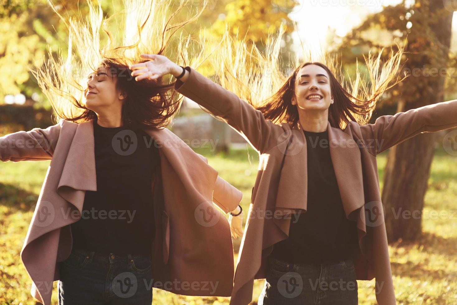 jonge gelukkige, lachende brunette tweelingmeisjes staan en spreiden de handen wijd open, hun haar is verdreven, gekleed in een casual jas in het zonnige herfstpark op de lichte wazige achtergrond. vrijheidsconcept foto