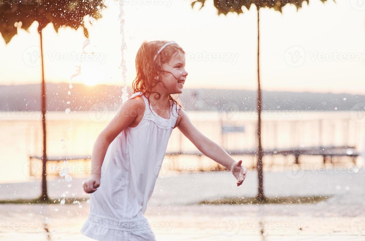 lachend en gelukkig. jong meisje spelen in de fontein op de zomerse hitte en het meer en de bossen achtergrond foto