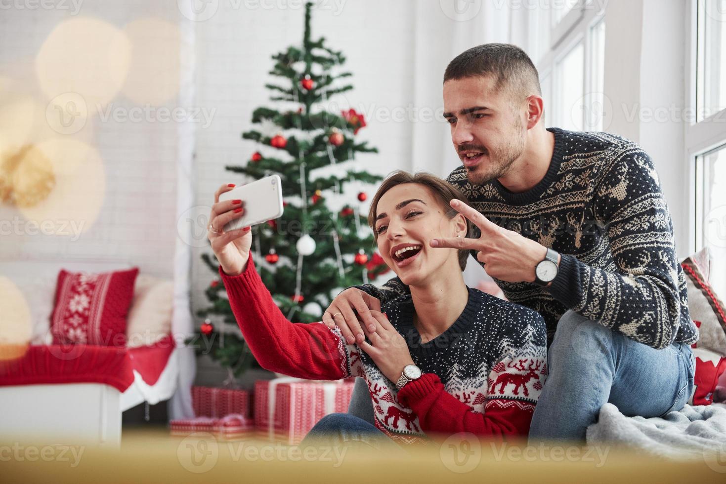 een selfie maken. kerel die gebaar van twee vingers toont. gelukkige jonge mensen zitten op de vensterbank in de kamer met kerstversieringen foto