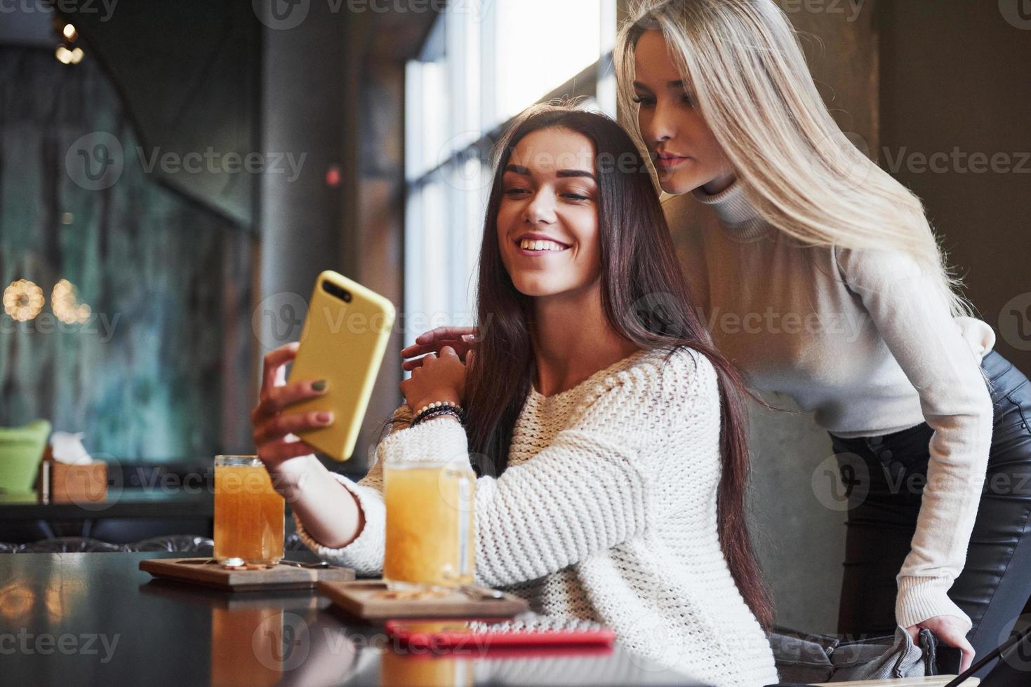 laten we een foto maken. in het restaurant. twee vriendinnen zitten binnen met gele drank en gebruiken de smartphone